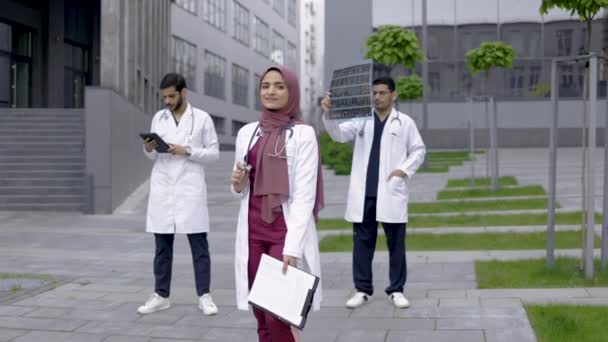 Médico musulmán con estetoscopio mira a la cámara de pie fuera del hospital frente a colegas varones — Vídeos de Stock