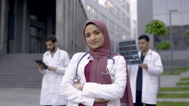 Dokter perempuan Arab mengenakan jilbab tersenyum sementara dua rekan laki-lakinya berdiri di belakang pintu — Stok Video