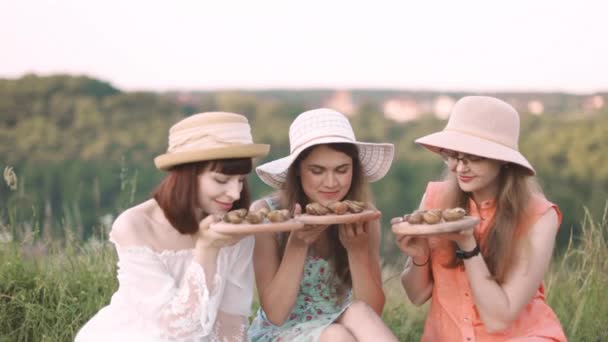 Vrouwelijke vrienden, picknick in een veld op een zonnige dag, ruikende geroosterde slakken op houten borden. — Stockvideo