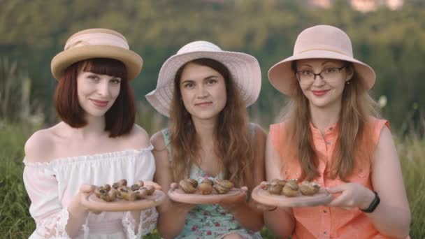 Красивые улыбающиеся девушки, сидящие на траве и демонстрирующие улитки на деревянных тарелках — стоковое видео