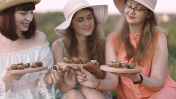 Три подруги, сидящие на одеяле на лугу и держащие деревянные тарелки с улитками — стоковое видео