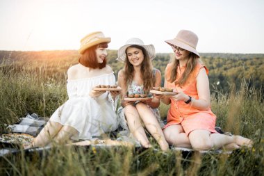 Güzel kadınlar, piknikte battaniyeye oturmuş, salyangozlu ahşap tabaklar tutuyorlar.