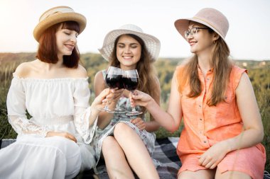 Üç genç kadın şapkalı ve elbiseli. Lezzetli yemeklerden, yaz pikniğinde şarap içiyorlar.