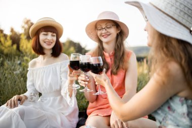 Üç güzel kadın, çimlerin üzerinde oturmuş piknik yaparken kırmızı şarap bardaklarını tokuşturuyor.