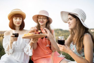 Üç genç bayan arkadaş gün batımında şarap, peynir ve meyvelerle piknik yapıyorlar.