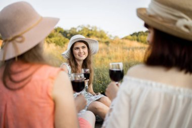 Şık şapkalı güzel bir kadın, elinde bir bardak kırmızı şarap, arkadaşlarıyla piknik yapıyor.