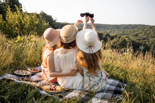 Vue arrière de jolies copines assises sur un plaid sur de l'herbe verte et levant les verres de vin rouge — Photo