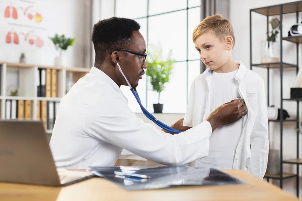 Médico afro-americano checando meninos batimento cardíaco e pulmões respiração durante sua visita ao centro médico. — Fotografia de Stock