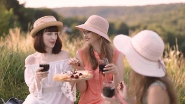 Üç genç bayan arkadaş gün batımında şarap, peynir ve meyvelerle piknik yapıyorlar.