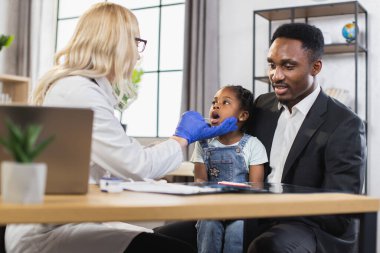 Kadın doktor genel teftiş sırasında küçük afro kızların boğazını spatulayla kontrol ediyor.