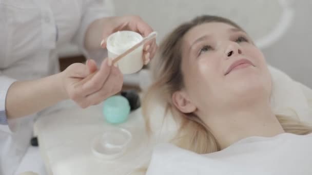 Close up de mãos esteticistas femininas, segurando frasco com creme, e aplicando-o usando espátula de madeira para rosto paciente na clínica de cosmetologia. Menina jovem com procedimentos de beleza no spa — Vídeo de Stock