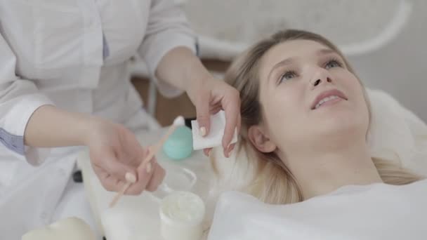 Zbliżenie żeńskich rąk kosmetyczki, trzymając słoik ze śmietanką i nakładając go za pomocą drewnianej szpachelki na twarz pacjenta w klinice kosmetologicznej. Młoda dziewczyna ma zabiegi kosmetyczne w spa — Wideo stockowe