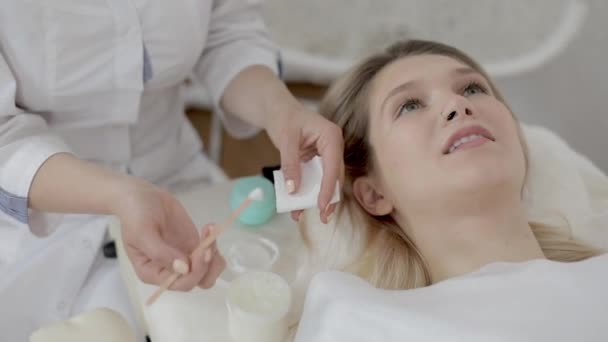 Close up of female beautician hands, holding jar with cream, and applying it using wooden spatula to patient face in cosmetology clinic. Young girl having beauty procedures in spa. — Stock Video