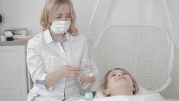 Bellissimo medico cosmetologo in camice bianco seduto dietro paziente femminile e applicare la crema sul viso. Giovane, bella donna sdraiata sul tavolo da massaggio e rilassante nel moderno centro cosmetologico. — Video Stock