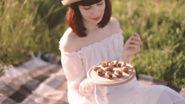 Молодая женщина сидит на траве и держит деревянные тарелки с вкусной французской едой — стоковое видео