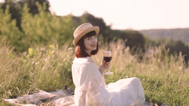 Young woman in summer dress and hat, posing and smiling at camera, holding glass with red wine — Stockvideo