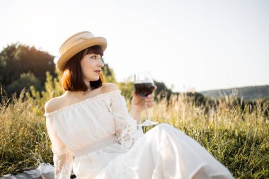 Beyaz elbiseli, hasır şapkalı, açık havada kırmızı şarap içen muhteşem kızıl saçlı kadının portresi.