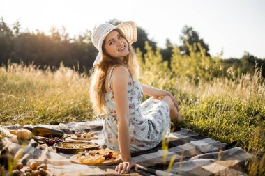 Doğa pikniğindeki kız lezzetli yiyeceklerle kareli battaniyenin üzerinde oturuyor.