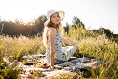 Doğa pikniğindeki kız lezzetli yiyeceklerle kareli battaniyenin üzerinde oturuyor.