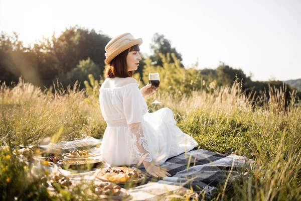 Mulher bonita em vestido branco e chapéu de palha desfrutando de belo pôr do sol de verão com vinho tinto ao ar livre — Fotografia de Stock