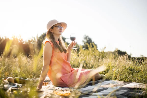 Jeune femme en robe et chapeau de paille boit du vin rouge sur le champ vert d'été — Photo