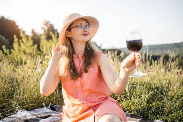 매력적 인 젊은 백인 아가씨가 여름 들판에서 붉은 포도주 잔으로 담요를 덮고 앉아 있는 모습 — 스톡 사진