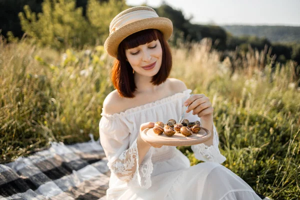 하얀 옷을 입고 밀짚모자를 쓰고 야외에서 피크닉에 앉아 맛있는 구운 달팽이를 먹는 여자 — 스톡 사진