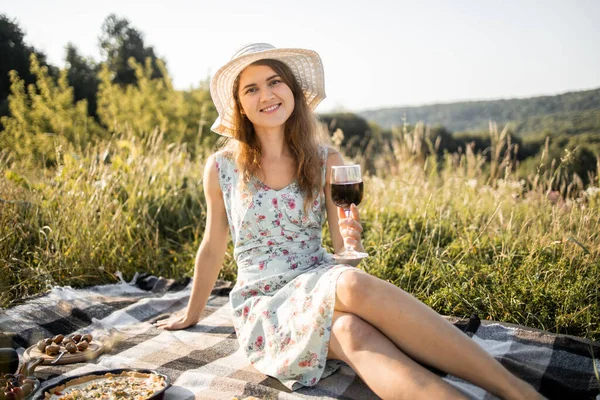 멋진 드레스와 모자를 입은 젊은 숙녀가 체크무늬 담요 위에 앉아 붉은 포도주를 마시고 있습니다 — 스톡 사진