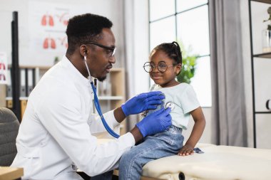 Afro-Amerikalı erkek doktor küçük Afrikalı kızları steteskop kullanarak kalp atışlarını ve nefeslerini kontrol ediyor.