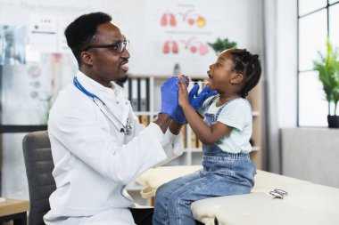 Siyah çocuk doktoru klinikte şirin bir çocuğun boğazını inceliyor.