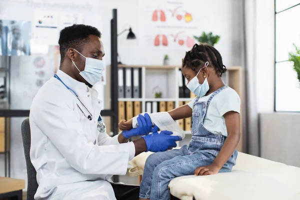 Médico Africano bandagem braço de menina de raça mista pouco na clínica moderna — Fotografia de Stock