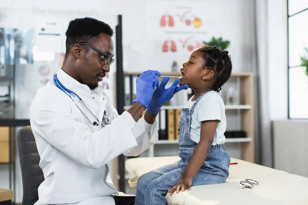 Afrikalı erkek doktor küçük kızın boğazını kontrol ediyor. Telifsiz Stok Imajlar