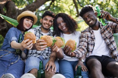 Doğada otururken ellerinde hamburger tutan çok ırklı arkadaşlar.