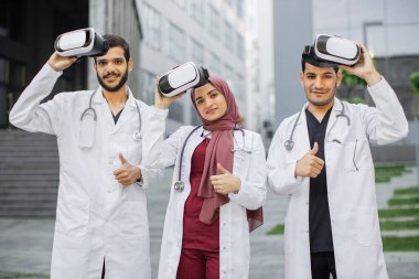 Sanal gerçeklik gözlüklü çok ırklı doktorlar hastanenin önünde baş parmağıyla poz veriyor.