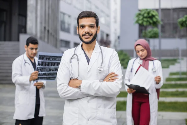Arabo o o indù uomo medico di fronte alla squadra multietnica, guardando la fotocamera con le braccia incrociate — Foto Stock