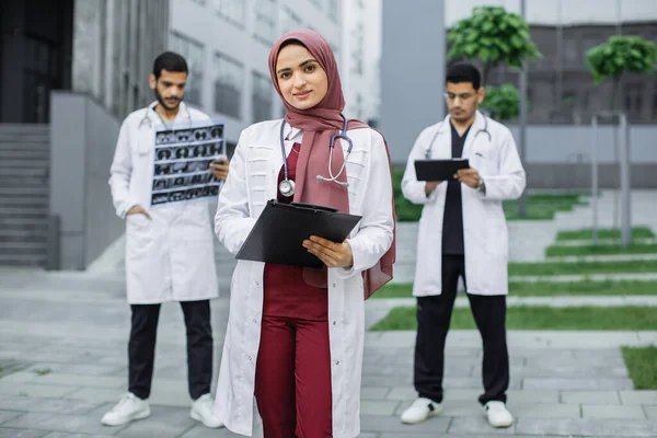 Muszlim kedves orvos hidzsábban, kint áll a kórház előtt, mögötte csapattal. — Stock Fotó
