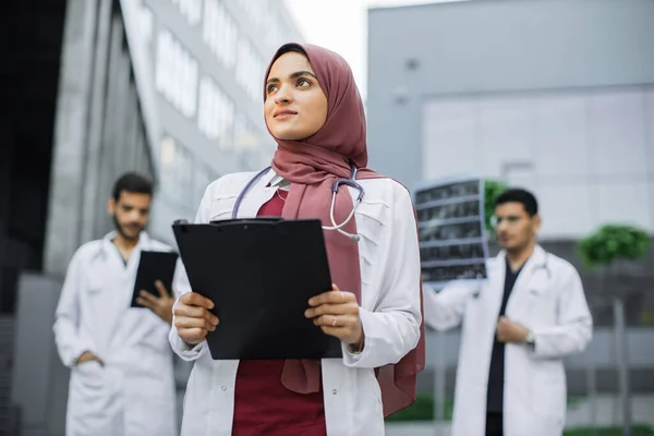 Mosolygó arab női orvos hidzsábban pózol a szabadban a modern klinika előtt, mögötte kollégákkal. — Stock Fotó