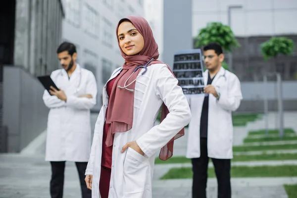男性同僚の前で病院の外に立っているカメラを見て聴診器を持つイスラム教徒の医師 — ストック写真