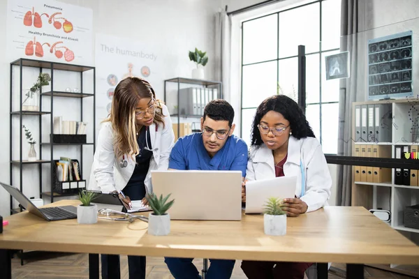 Drei multiethnische Ärzte, die zusammen mit Laptop arbeiten, während sie in einer modernen Klinik am Tisch sitzen — Stockfoto
