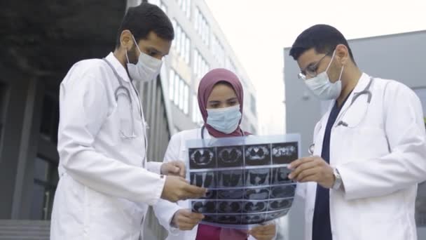 Drie artsen neurologen, dragen uniform en masker, poseren buiten en kijken naar MRI-scan. — Stockvideo