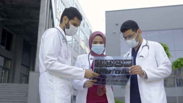 Team van drie jonge diverse experts artsen kijken naar MRI foto in de voorkant van kliniek buiten — Stockvideo