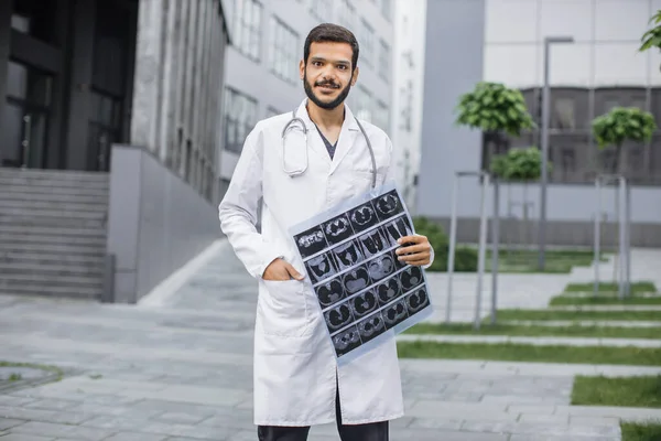 Male Arab doctor smiling at camera, holding tomography scan, standing outdoors near clinic. — Stock Photo, Image