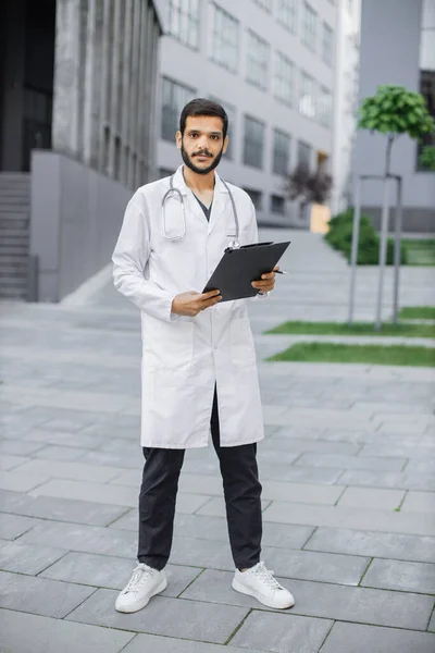 Arabian healthcare worker in white coat, standing with clipboard folder in hands outdoors — Stock Photo, Image