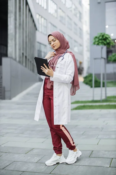 Muslim doctor in hijab, working with patients records on digital tablet standing outside clinic — Stock Photo, Image