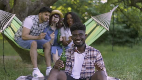 Черный человек пьет и ест, пока друзья разговаривают на открытом воздухе — стоковое видео
