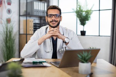 Beyaz önlüklü ve gözlüklü erkek Arap doktor, elinde laptop ve kağıtlarla masada oturuyor.