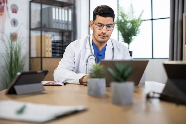 Professionnel jeune spécialiste médical arabe en utilisant un ordinateur portable au travail dans une clinique moderne — Photo