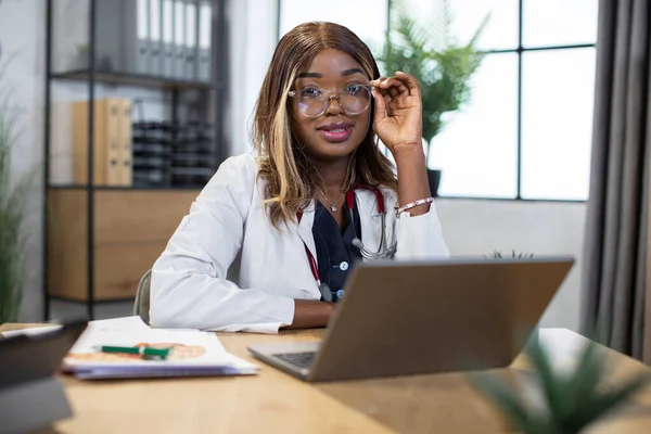 Afro-Amerikaanse arts vrouw, zitten aan de tafel in de moderne kliniek, werken aan laptop pc — Stockfoto