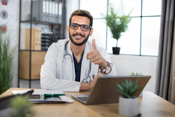 Medico arabo in camice bianco, seduto nello studio medico alla scrivania con computer portatile, mostrando il pollice in alto — Foto Stock