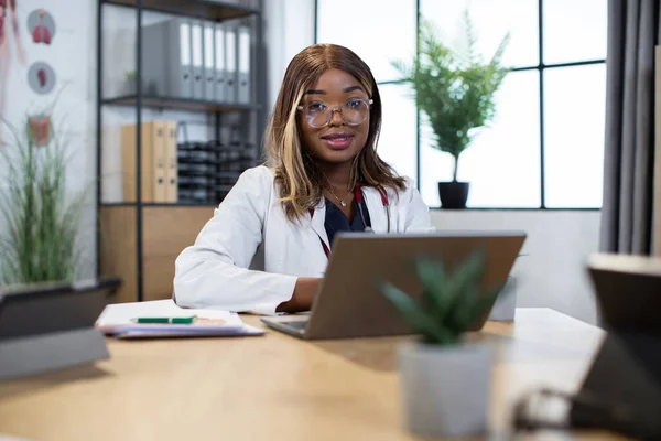 女性のアフリカ人医師はカメラを見ながらテーブルに座りノートパソコンを使い ストックフォト